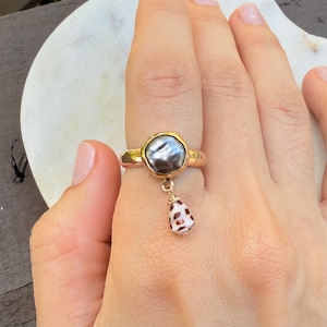 May include: A gold ring featuring a central dark gray, iridescent, irregular-shaped stone set in a bezel. A small, teardrop-shaped white shell with brown spots hangs from the base of the stone setting by a thin gold wire loop.