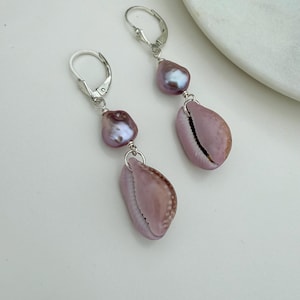 May include: A pair of dangle earrings featuring a light purple cowrie shell and a light purple pearl. The earrings have a silver-colored hook closure. The shells have a natural, textured appearance, and the pearls have a subtle iridescent sheen.
