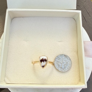 May include: A gold ring with a triangular, cream-colored stone with brown markings. The ring is displayed in a white box next to a dime for size comparison. The ring's band is thin and delicate.