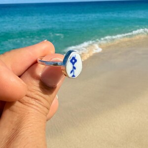 May include: A silver ring featuring a smooth, oval-shaped white ceramic inlay. The inlay is decorated with a small, hand-painted blue flower design with a single stem and leaves. The metal band is polished and reflects light, creating a bright appearance.