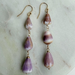 May include: A pair of dangle earrings featuring three teardrop-shaped beads in shades of purple, white, and brown. The beads are connected by gold-colored wire and hang from gold-colored ear wires. The earrings are displayed on a light-colored surface.