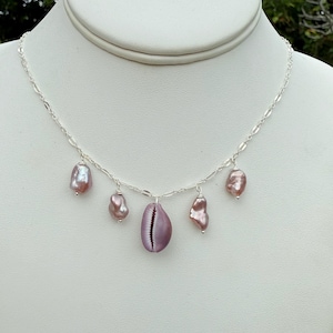 May include: A silver necklace featuring a central lavender-colored seashell pendant, surrounded by four irregularly shaped, mauve-colored pearls. The necklace is displayed on a white mannequin bust against a blurred green background.