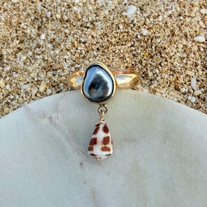May include: A gold ring featuring a dark gray, irregular-shaped pearl set in a bezel, with a small, cone-shaped seashell charm with brown and white markings dangling from a gold wire loop attached to the base of the pearl setting.