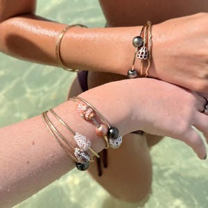May include: Gold-toned bangles and bracelets adorn wrists in a sunlit setting. Some bracelets feature natural seashell and pearl accents. The jewelry is worn in a body of water, suggesting a beach or aquatic theme.