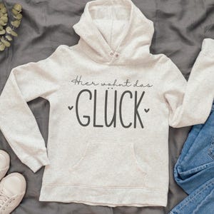 K&ouml;nnte beinhalten: Ein hellgrauer Kapuzenpullover mit Fronttasche und Kapuze. Der Kapuzenpullover tr&auml;gt den Text "Hier wohnt das GL&Uuml;CK" in Dunkelgrau mit kleinen Herzsymbolen. Der Kapuzenpullover wird mit einer blauen Jeans und wei&szlig;en Turnschuhen kombiniert.