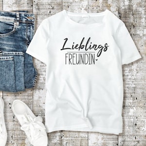 K&ouml;nnte beinhalten: Wei&szlig;es T-Shirt mit dem Text "Lieblings FREUNDIN" in schwarzer Schrift. Das Shirt liegt neben einer Jeans und wei&szlig;en Turnschuhen auf einer hellen Holzoberfl&auml;che.