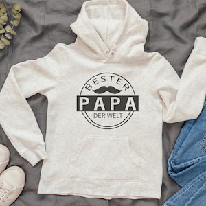 May include: A light gray hoodie with a front pocket and a hood. The hoodie features a black graphic that says "BESTER PAPA DER WELT" with a mustache illustration. The hoodie is paired with blue jeans and white sneakers.