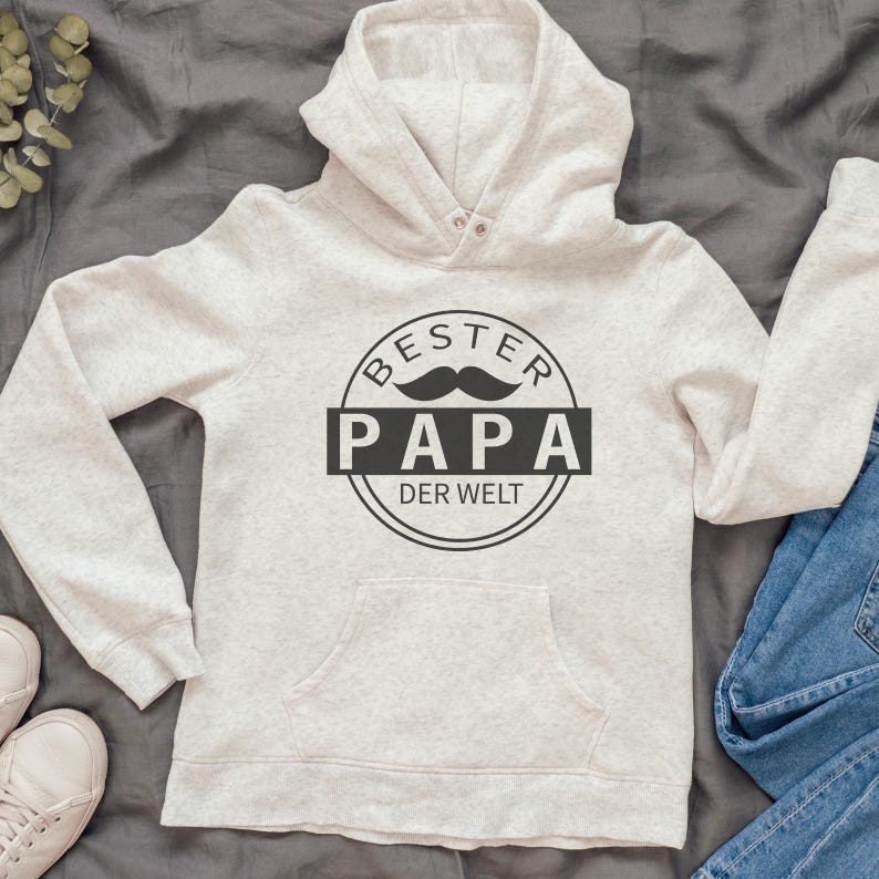 May include: A light gray hoodie with a front pocket and a hood. The hoodie features a black graphic that says "BESTER PAPA DER WELT" with a mustache illustration. The hoodie is paired with blue jeans and white sneakers.