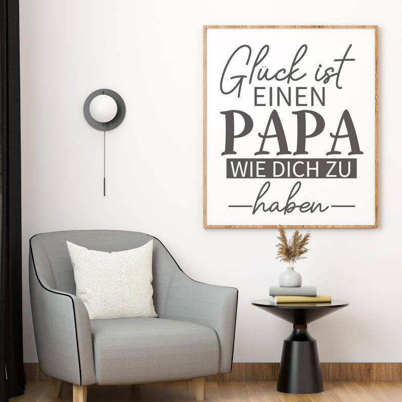 May include: A framed wall art print with the text "Gl&uuml;ck ist einen PAPA wie dich zu haben" in black lettering. The artwork hangs above a gray armchair with a white pillow. A small side table holds books and a vase.