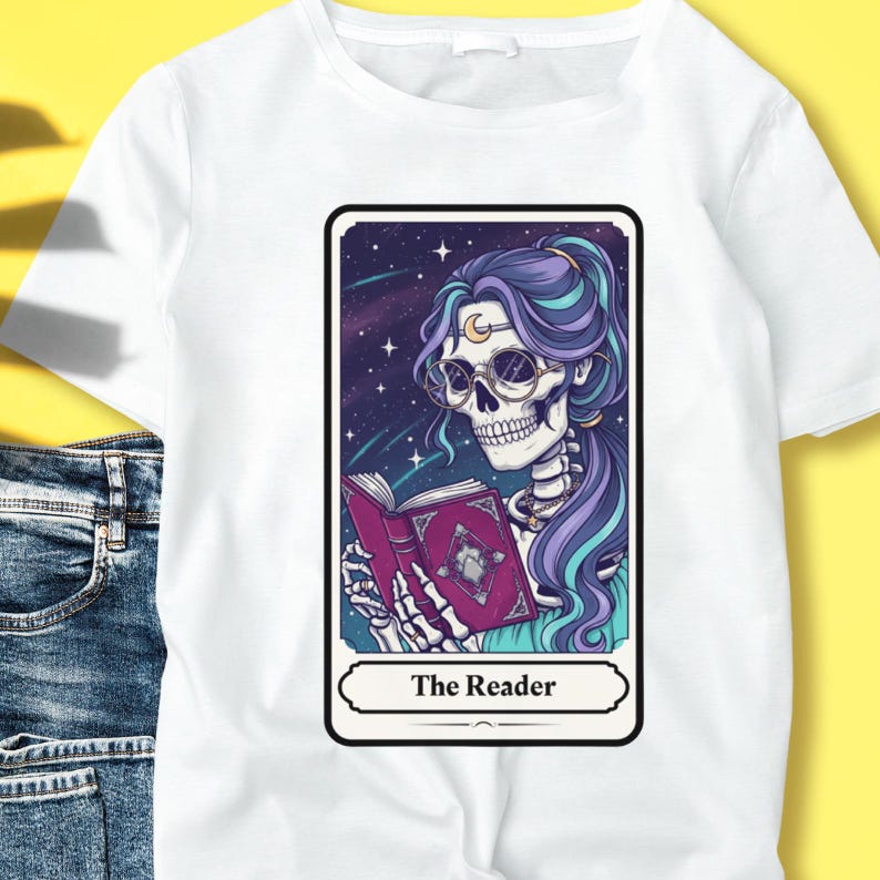 Puede incluir: Camiseta blanca con un dise&ntilde;o de carta del tarot. La carta representa un esqueleto con pelo morado y gafas leyendo un libro sobre un fondo estrellado. La carta est&aacute; etiquetada como "The Reader".