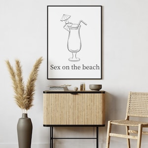 May include: Black and white framed print featuring a cocktail illustration with a straw and umbrella garnish. The text "Sex on the beach" is below the image. The print is displayed above a wooden cabinet.