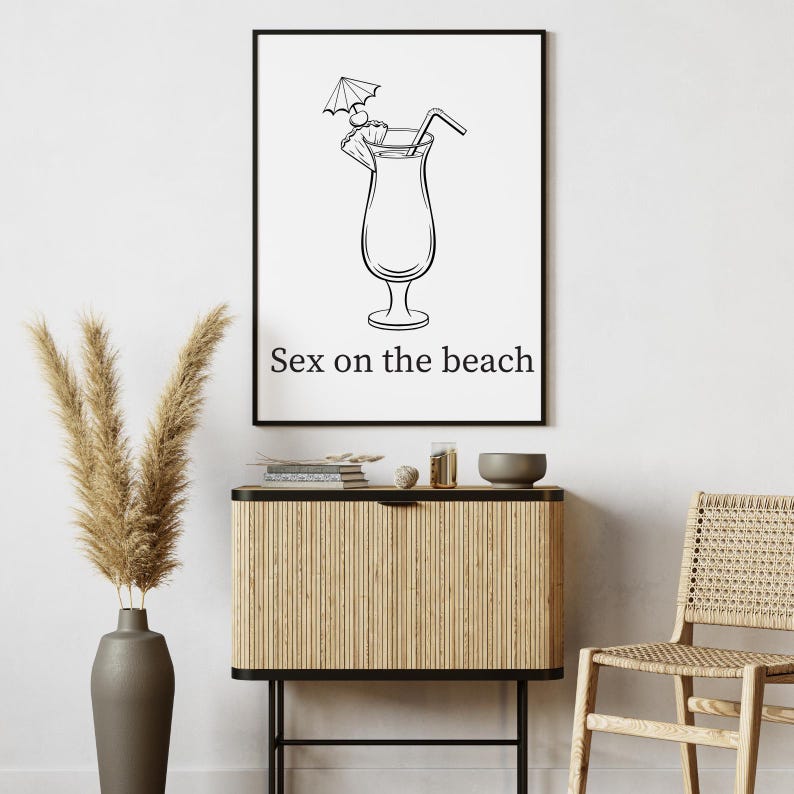 May include: Black and white framed print featuring a cocktail illustration with a straw and umbrella garnish. The text "Sex on the beach" is below the image. The print is displayed above a wooden cabinet.