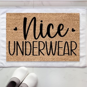 May include: A rectangular coir doormat with the words "Nice UNDERWEAR" in black, cursive font. Two small black hearts are on either side of "Nice". The mat sits on a white towel, with white slippers visible in the foreground.