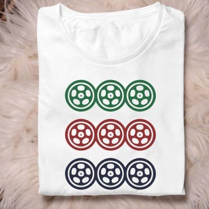 May include: White t-shirt with a design of three rows of three stylized wheels. The top row is green, the middle row is red, and the bottom row is navy blue. The wheels have a central design with six holes.