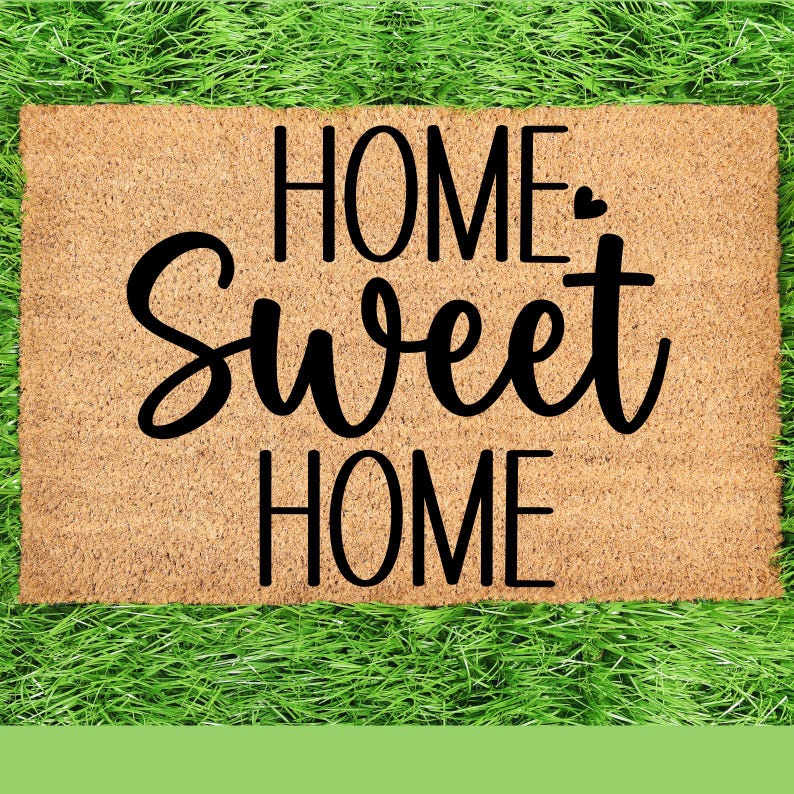May include: A rectangular doormat made of natural coir fibre. The mat features the words "HOME Sweet HOME" in a black, cursive font. The doormat is placed on a bed of green grass.