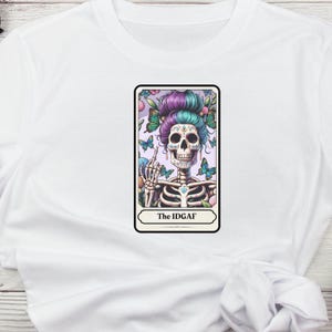 Puede incluir: Camiseta blanca con un gr&aacute;fico colorido de un esqueleto con un tocado floral y mariposas. El esqueleto est&aacute; mostrando el dedo medio. El texto "The IDGAF" se muestra debajo de la imagen.