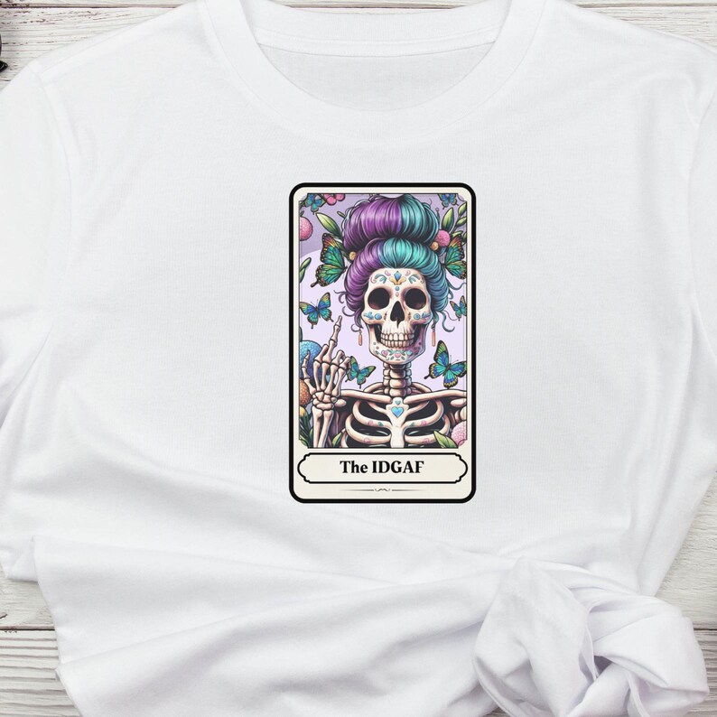 Puede incluir: Camiseta blanca con un gr&aacute;fico colorido de un esqueleto con un tocado floral y mariposas. El esqueleto est&aacute; mostrando el dedo medio. El texto "The IDGAF" se muestra debajo de la imagen.