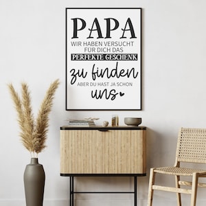 May include: A framed black and white print with the word "PAPA" at the top. The text below reads "WIR HABEN VERSUCHT F&Uuml;R DICH DAS PERFEKTE GESCHENK zu finden ABER DU HAST JA SCHON uns". The print is displayed above a wooden cabinet.