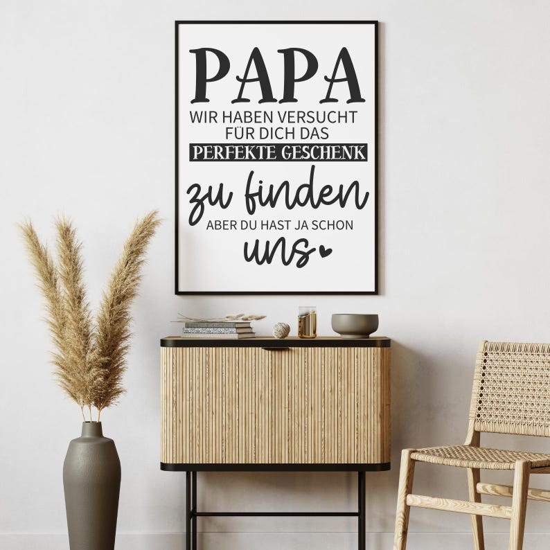 May include: A framed black and white print with the word "PAPA" at the top. The text below reads "WIR HABEN VERSUCHT F&Uuml;R DICH DAS PERFEKTE GESCHENK zu finden ABER DU HAST JA SCHON uns". The print is displayed above a wooden cabinet.