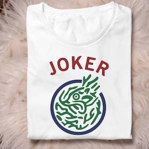 May include: White t-shirt featuring the word "JOKER" in red. Below the word is a blue circle containing a green and white graphic. The shirt is folded.