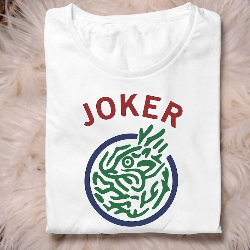 May include: White t-shirt featuring the word "JOKER" in red. Below the word is a blue circle containing a green and white graphic. The shirt is folded.