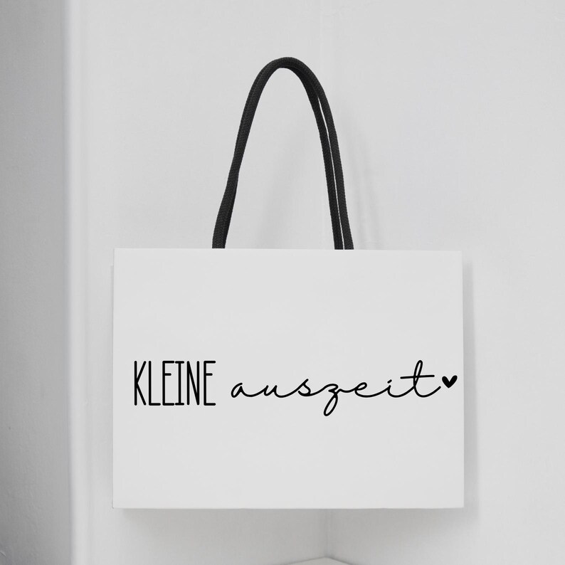 K&ouml;nnte beinhalten: Wei&szlig;e Tragetasche mit schwarzen Henkeln. Auf der Tasche steht in schwarzer Schrift "KLEINE auszeit", am Ende ein kleines Herzsymbol. Die Tasche h&auml;ngt vor einer wei&szlig;en Wand.