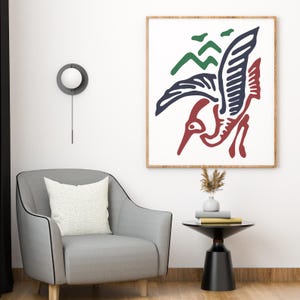 May include: A framed art print with a bird design in red, navy blue, and green. The artwork is set against a white background within a wooden frame. The print hangs above a grey armchair and a small black side table, creating a modern aesthetic.