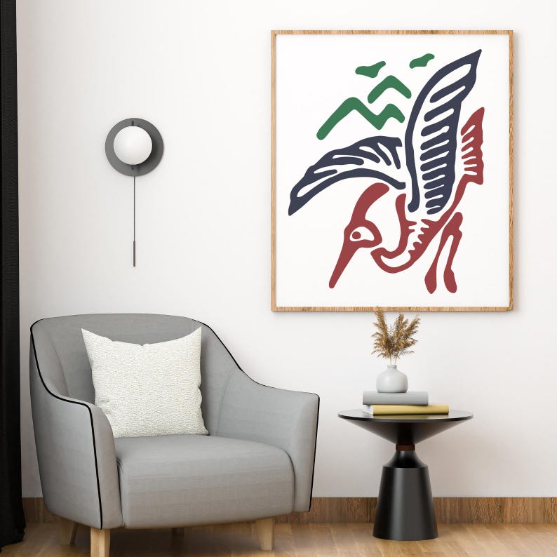 May include: A framed art print with a bird design in red, navy blue, and green. The artwork is set against a white background within a wooden frame. The print hangs above a grey armchair and a small black side table, creating a modern aesthetic.