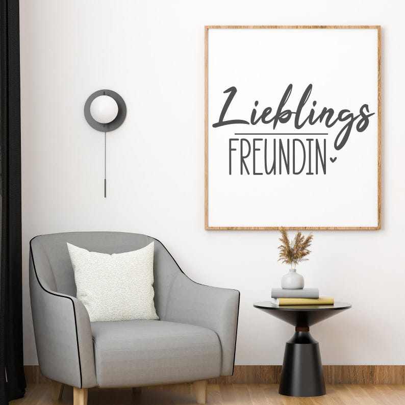 K&ouml;nnte beinhalten: Ein gerahmter Druck mit den Worten "Lieblings FREUNDIN" in schwarzer Schrift und einem Herz. Der Druck h&auml;ngt &uuml;ber einem grauen Sessel mit einem wei&szlig;en Kissen. Ein kleiner schwarzer Beistelltisch enth&auml;lt B&uuml;cher und eine Vase mit getrockneten Blumen. Eine moderne Wanduhr ist ebenfalls sichtbar.