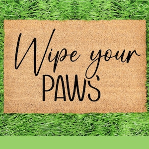 May include: A rectangular doormat made of natural coir fibre, with the words "Wipe your PAWS" in a black, cursive font. The mat is set against a background of vibrant green artificial grass.