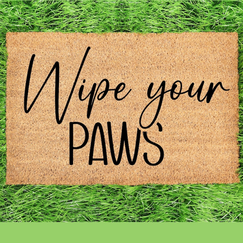 May include: A rectangular doormat made of natural coir fibre, with the words "Wipe your PAWS" in a black, cursive font. The mat is set against a background of vibrant green artificial grass.