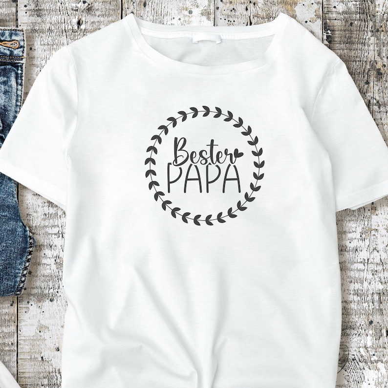 May include: White t-shirt with a crew neck featuring the text "Bester PAPA" in black script inside a black leaf wreath. The shirt is made of a soft, comfortable material.