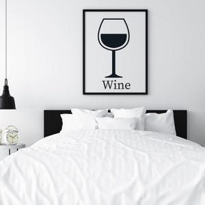 May include: A framed black and white print of a wine glass with the word "Wine" below it, hanging above a bed with white bedding. A black lamp and a small alarm clock are visible.