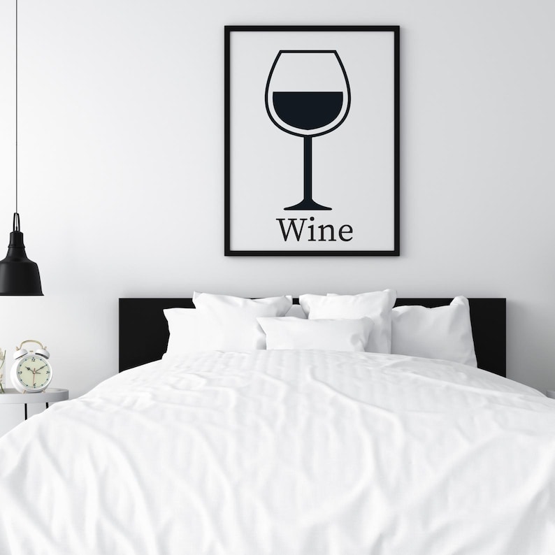 May include: A framed black and white print of a wine glass with the word "Wine" below it, hanging above a bed with white bedding. A black lamp and a small alarm clock are visible.