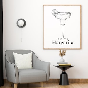 May include: A framed black and white illustration of a margarita cocktail with a lime slice. The word "Margarita" is printed below the glass. The artwork is displayed on a white wall above a gray armchair and a small black side table.