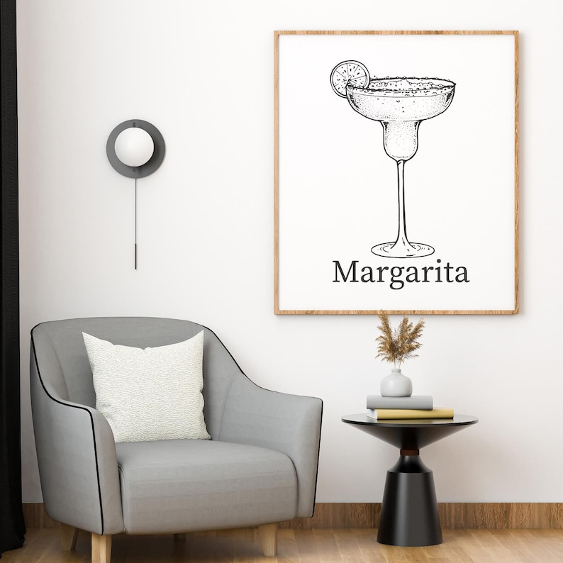May include: A framed black and white illustration of a margarita cocktail with a lime slice. The word "Margarita" is printed below the glass. The artwork is displayed on a white wall above a gray armchair and a small black side table.