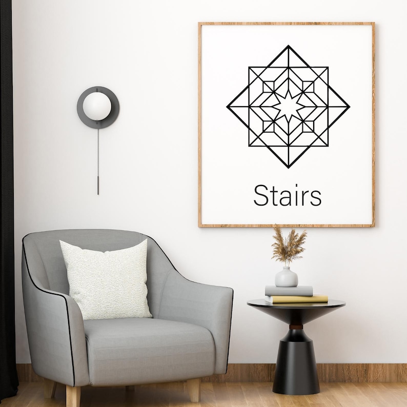 May include: A framed print with a geometric black and white design and the word "Stairs" below it hangs on a white wall. A gray armchair with a white pillow sits in front of the wall. A small black table holds books and a vase.