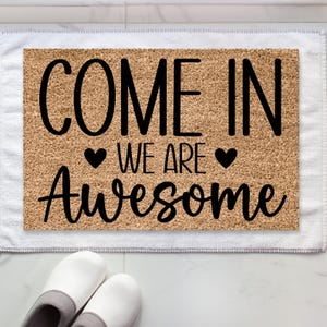 May include: A rectangular doormat with the words "COME IN WE ARE AWESOME" in black script. The mat is made of a light brown, textured material and is placed on a white towel. Two pairs of white slippers are at the bottom of the image.