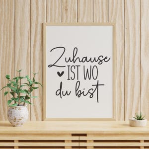 K&ouml;nnte beinhalten: Ein gerahmter Druck mit dem deutschen Satz "Zuhause ist wo du bist" in schwarzer Schrift. Der Druck befindet sich in einem hellen Holzrahmen, der auf einer Holzoberfl&auml;che mit einer kleinen Topfpflanze und einer dekorativen Vase mit einer Pflanze steht.