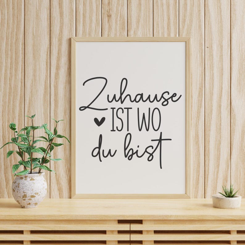 K&ouml;nnte beinhalten: Ein gerahmter Druck mit dem deutschen Satz "Zuhause ist wo du bist" in schwarzer Schrift. Der Druck befindet sich in einem hellen Holzrahmen, der auf einer Holzoberfl&auml;che mit einer kleinen Topfpflanze und einer dekorativen Vase mit einer Pflanze steht.