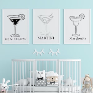 May include: Three framed black and white cocktail prints: Cosmopolitan, Martini, and Margherita. The prints are displayed above a white crib in a nursery with a light blue wall. Decorative pillows and a doll are in the crib.