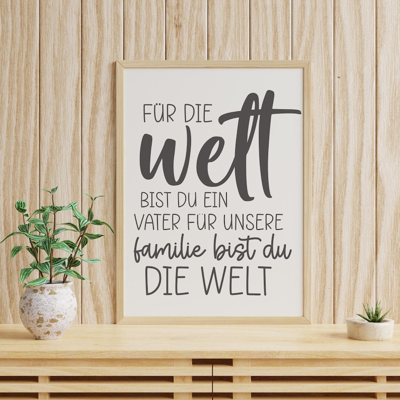 May include: A framed print with the words "F&Uuml;R DIE Welt BIST DU EIN VATER F&Uuml;R UNSERE familie BIST DU DIE WELT" in black script. The print is in a light wood frame, set on a wooden surface with a small plant and a decorative vase.