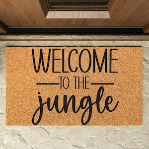 May include: A rectangular doormat made of natural coir fibre. The mat features the phrase "WELCOME TO THE JUNGLE" in black, stylised lettering. The mat is placed in front of a wooden door.