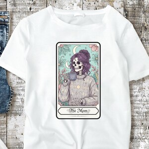 Puede incluir: Camiseta blanca con un dise&ntilde;o de carta del tarot. La carta representa una figura de esqueleto con pelo morado, sosteniendo una taza y vistiendo un su&eacute;ter. La carta est&aacute; enmarcada con elementos florales y el texto "The Mom".