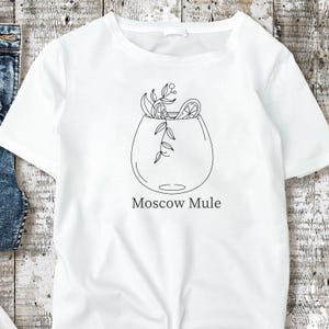 May include: White t-shirt featuring a black line drawing of a Moscow Mule cocktail. The illustration includes a glass with garnishes and the words "Moscow Mule" in a simple font. The design is centered on the shirt.