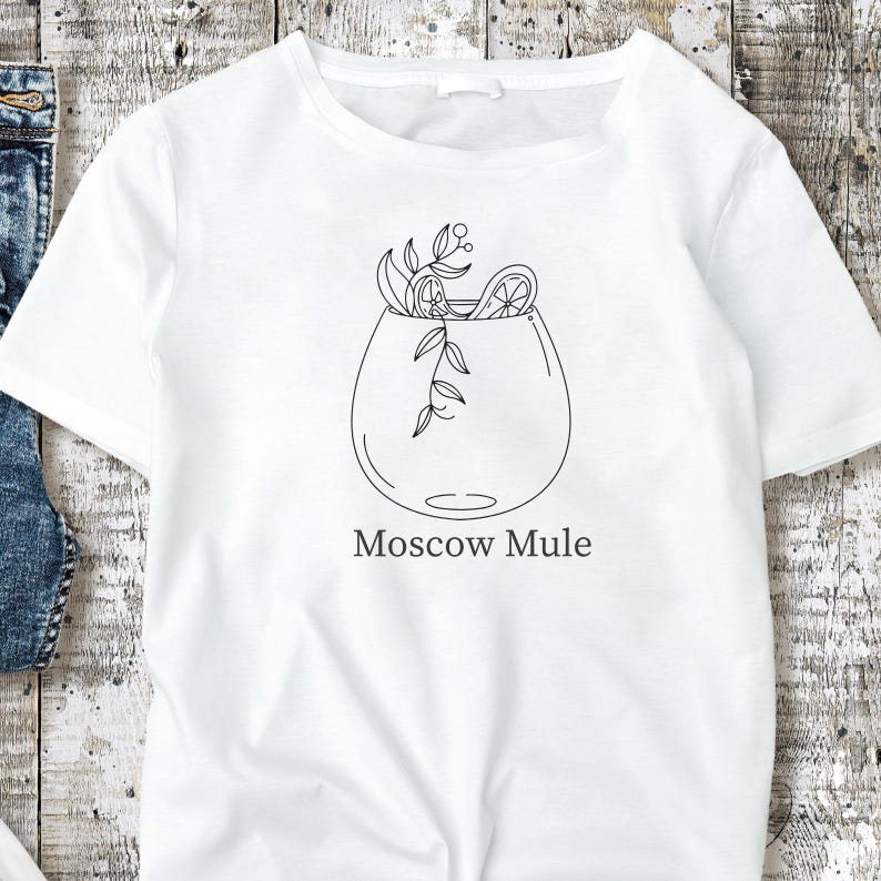May include: White t-shirt featuring a black line drawing of a Moscow Mule cocktail. The illustration includes a glass with garnishes and the words "Moscow Mule" in a simple font. The design is centered on the shirt.