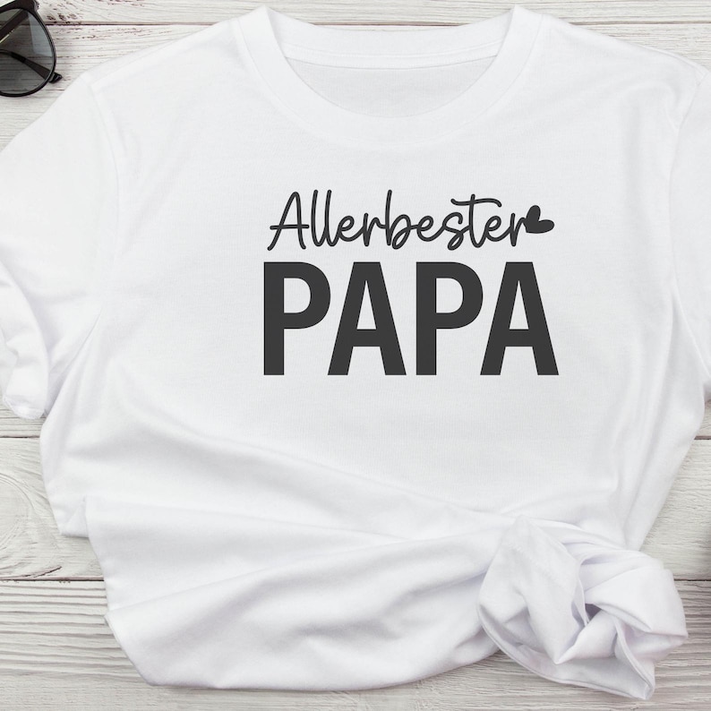 May include: White t-shirt with the German text "Allerbester PAPA" in black, with a small heart above the word "PAPA". The shirt is made of a soft material and has a crew neck.