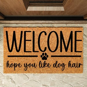 May include: A rectangular doormat with the word "WELCOME" in large black letters. Below, it reads "hope you like dog hair" in a script font, with a paw print symbol. The mat is brown and sits on a stone surface.