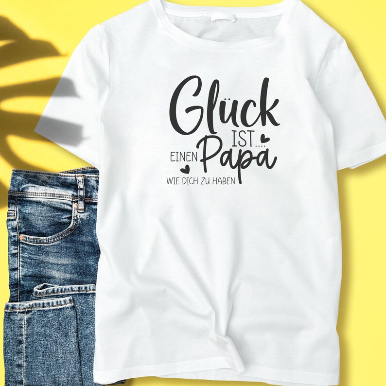 May include: White t-shirt with the text "Gl&uuml;ck ist einen Papa wie dich zu haben" in black script. The shirt is paired with blue denim jeans. The text translates to "Happiness is having a dad like you."