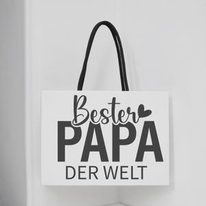 May include: A white wooden sign with the words "Bester PAPA DER WELT" in black lettering, with a black rope handle. The sign is hanging on a white wall.